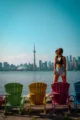 Portrait d'une femme debout sur une chaise Muskoka colorée (rouge) sur les îles de Toronto, avec une vue panoramique complète sur la skyline de Toronto et la Tour CN derrière elle, sous un ciel bleu.