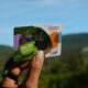Main tenant la carte Pass Ardèche 3 jours devant un paysage naturel sous un ciel bleu.